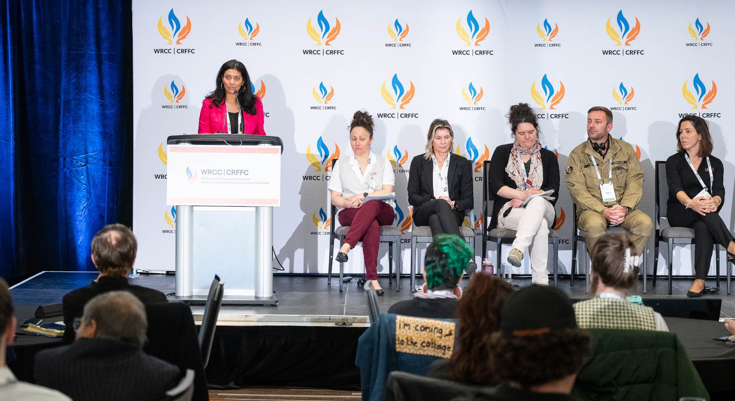 A speaker at a podium addresses the audience beside a seated panel at a WRCC event.